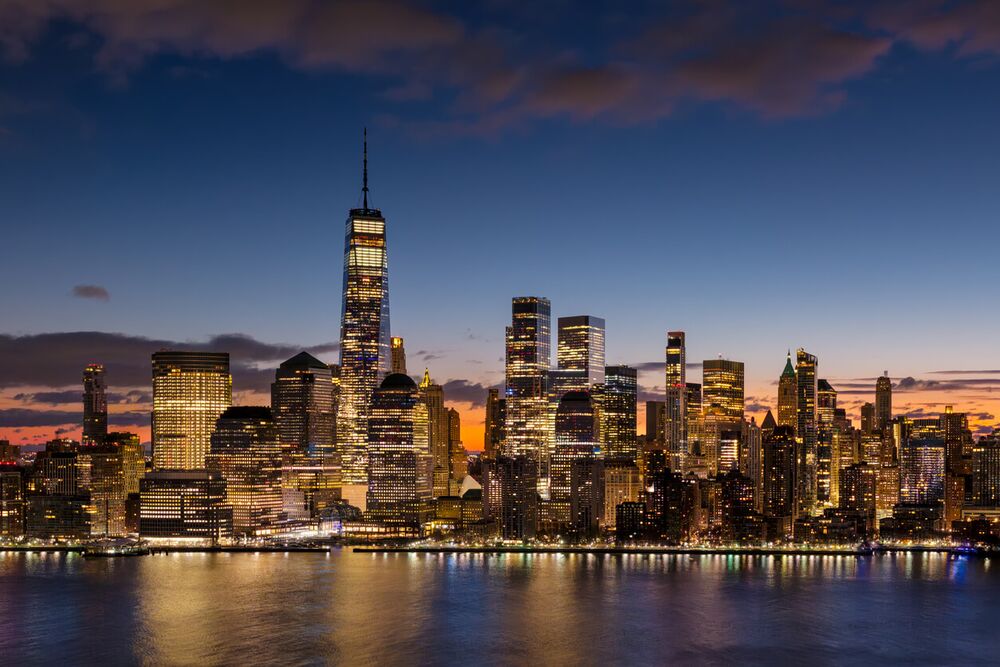 The Lower Manhattan Financial District skyline at dusk, with One World Trade Center lit against the sky and reflections across the water